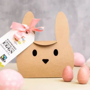 White Chocolate Raspberry Mini Eggs unboxed sitting next to White Chocolate Raspberry Mini Eggs in a rabbit themed box sitting on a table with coloured pink eggs around it.